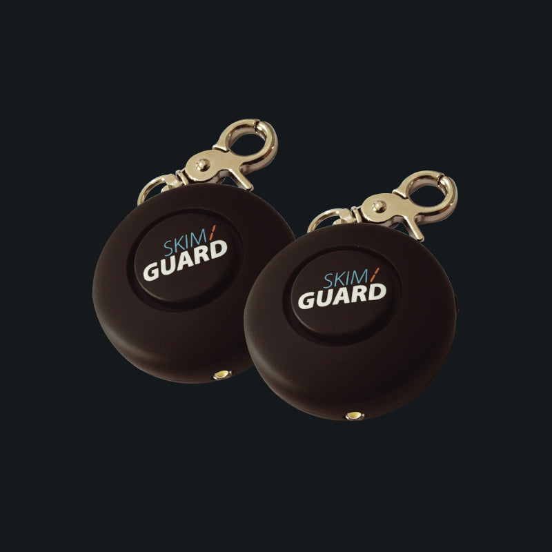 Two Skimguard Personal Alarms with front buttons and attached metal clips, shown overlapping.