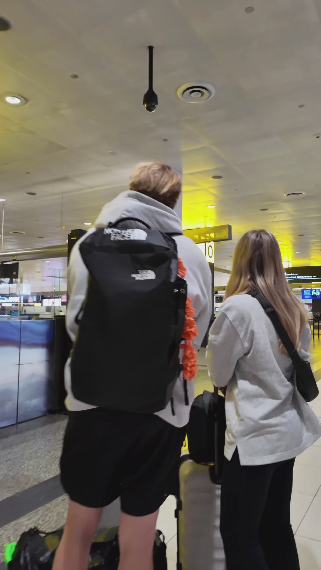 Two people standing in an airport terminal with luggage, viewed from behind.